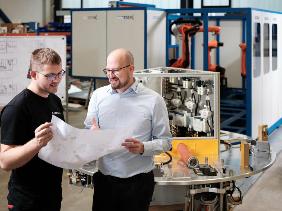 Zwei Mitarbeiter der Firma Gatzsch Schweißtechnik stehen in einer Produktionshalle vor einer Schweißanlage und betrachten gemeinsam technische Zeichnungen. Der Mitarbeiter auf der linken Seite trägt ein schwarzes T-Shirt mit dem Logo von Gatzsch, während der Mitarbeiter rechts ein Hemd mit Gatzsch-Logo trägt. Im Hintergrund sind weitere Maschinen und technische Anlagen zu sehen. Das Bild symbolisiert die enge Zusammenarbeit und den Austausch zwischen Entwicklung und Produktion bei der Konstruktion und Optimierung von Schweißanlagen.