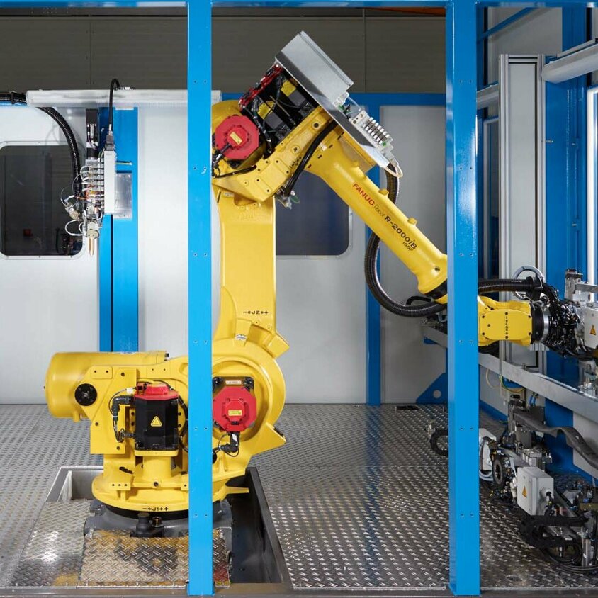 Eine industrielle Klebeanlage mit einem gelben FANUC-Roboterarm in einem abgeschirmten Arbeitsbereich. Der Roboter ist in der Mitte der Anlage positioniert, bereit für automatisierte Aufgaben. Umgeben von blauen Rahmenstrukturen, zeigt die Szene eine hochmoderne Fertigungsumgebung mit mechanischen Komponenten und Steuergeräten. Diese Klebeanlage steht für Präzision, Effizienz und Automatisierung in der industriellen Fügetechnik, wie sie bei Gatzsch Schweißtechnik eingesetzt wird.