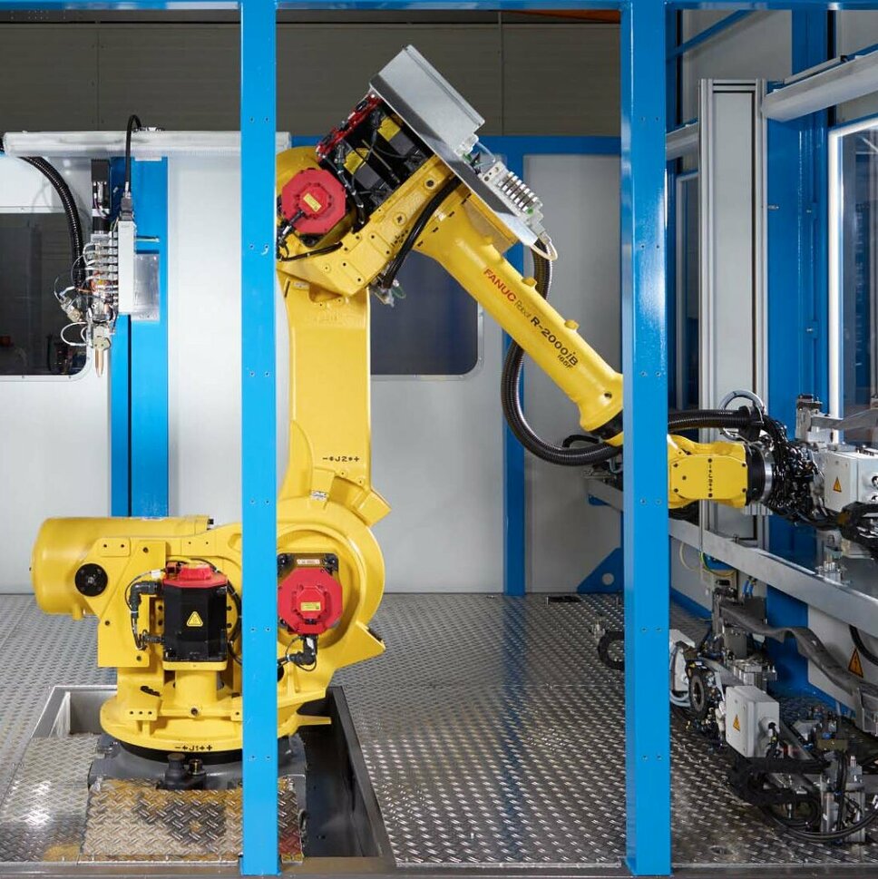 Eine industrielle Klebeanlage mit einem gelben FANUC-Roboterarm in einem abgeschirmten Arbeitsbereich. Der Roboter ist in der Mitte der Anlage positioniert, bereit für automatisierte Aufgaben. Umgeben von blauen Rahmenstrukturen, zeigt die Szene eine hochmoderne Fertigungsumgebung mit mechanischen Komponenten und Steuergeräten. Diese Klebeanlage steht für Präzision, Effizienz und Automatisierung in der Produktion, wie sie bei Gatzsch Schweißtechnik eingesetzt wird.