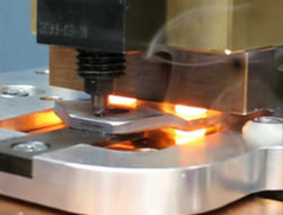 Nahaufnahme eines industriellen Warmnietvorgangs. Ein erhitztes Nietwerkzeug presst ein glühendes Metallteil auf ein Werkstück, um eine sichere Verbindung herzustellen. Der Bereich um das Werkzeug leuchtet aufgrund der Hitzeeinwirkung, während leichter Rauch aufsteigt. Diese Szene zeigt die Präzision und Effizienz des Warmnietens, eine Technologie, die in der Fertigung eingesetzt wird.