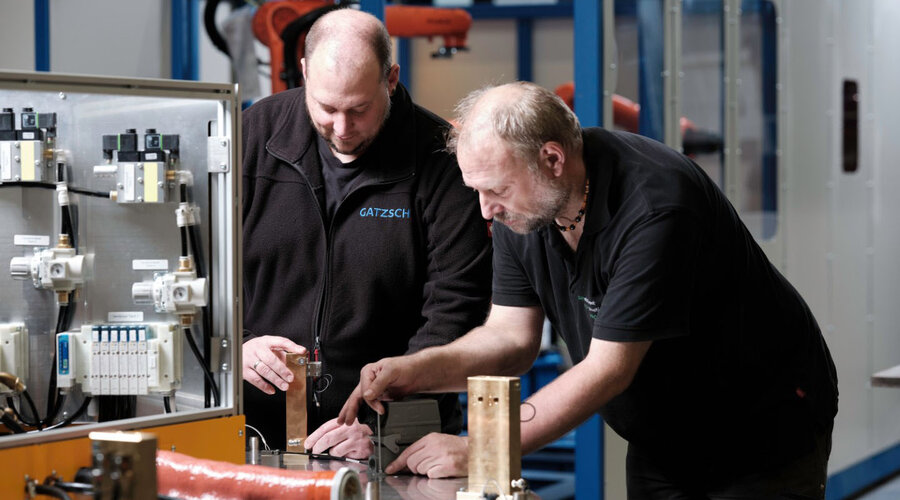 Zwei Mitarbeiter der Firma Gatzsch Schweißtechnik arbeiten gemeinsam an einer Schweißanlage in der Anlagenmontage. Sie überprüfen und diskutieren die Bauteile einer Schweißanlage, wobei einer der Mitarbeiter auf ein spezielles Element zeigt. Im Hintergrund ist eine moderne Produktionshalle mit weiteren Maschinen zu erkennen. Das Bild vermittelt die Zusammenarbeit und das Fachwissen, das für die Inbetriebnahme und Wartung komplexer Schweißanlagen erforderlich ist.