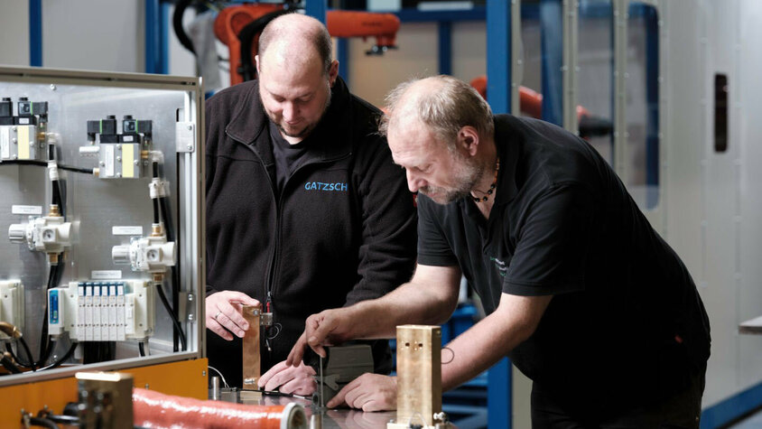 Zwei Mitarbeiter der Firma Gatzsch Schweißtechnik arbeiten gemeinsam an einer Schweißanlage in der Anlagenmontage. Sie überprüfen und diskutieren die Bauteile einer Schweißanlage, wobei einer der Mitarbeiter auf ein spezielles Element zeigt. Im Hintergrund ist eine moderne Produktionshalle mit weiteren Maschinen zu erkennen. Das Bild vermittelt die Zusammenarbeit und das Fachwissen, das für die Inbetriebnahme und Wartung komplexer Schweißanlagen erforderlich ist.