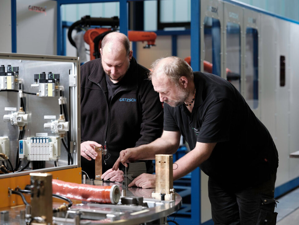 wo employees of Gatzsch Schweißtechnik work together on a welding system during the assembly process. They are reviewing and discussing the components of the welding system, with one employee pointing to a specific element. In the background, a modern production hall with more machines can be seen. The image conveys the collaboration and expertise required for the commissioning and maintenance of complex welding systems.