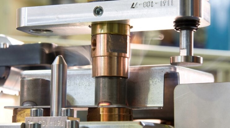 Detailaufnahme einer Buckelschweißanlage während des Schweißvorgangs. Zu sehen sind Metallkomponenten und Elektroden, die für das präzise Buckelschweißen positioniert sind. Das Bild zeigt die Feinmechanik der Schweißvorrichtung der Firma Gatzsch Schweißtechnik.