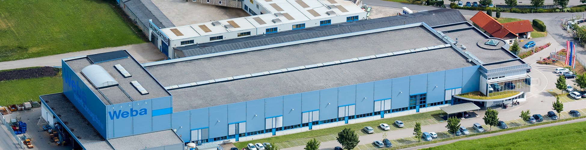 Die Luftaufnahme zeigt das beeindruckende Headquarters und Entwicklungszentrum von weba in Dietach. Die Produktionsstätte erstreckt sich über das Gelände, während im Vordergrund die Pressenhalle mit der deutlich sichtbaren Beschriftung "WEBA" zu sehen ist. Die Architektur wirkt modern und funktional, während die Umgebung von Grünflächen und Straßen umgeben ist. Die Szene vermittelt einen Eindruck von Größe und Professionalität, während das "WEBA"-Logo die Identität des Unternehmens betont.